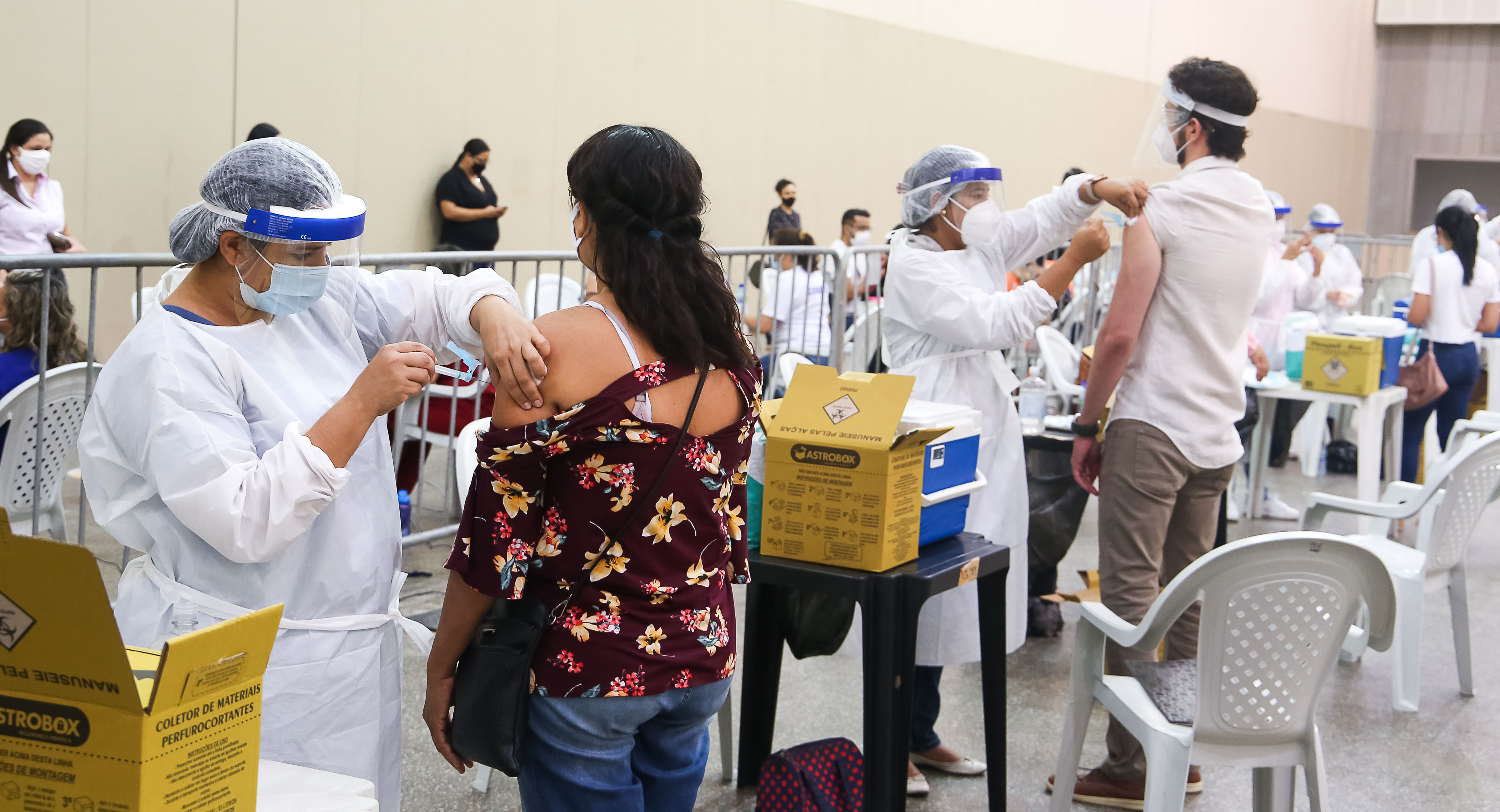vacinadores aplicando vacina em uma mulher e um homem de costas no Centro de Eventos e fila de pessoas ao fundo aguardando